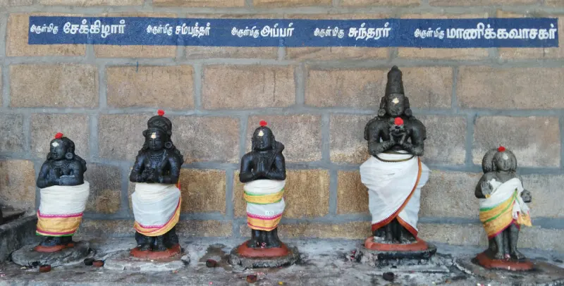 Five small black stone figures with folded hands, wearing white cloth, below a blue Tamil name board