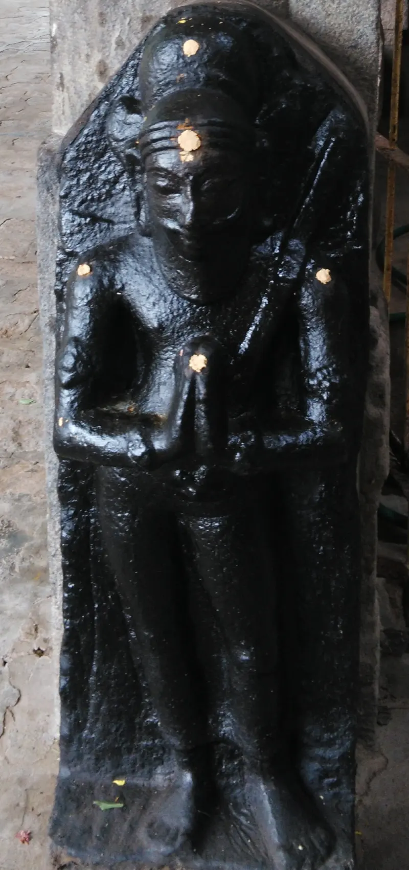 A black stone relief of a standing figure with folded hands, with dots of gold on its forehead