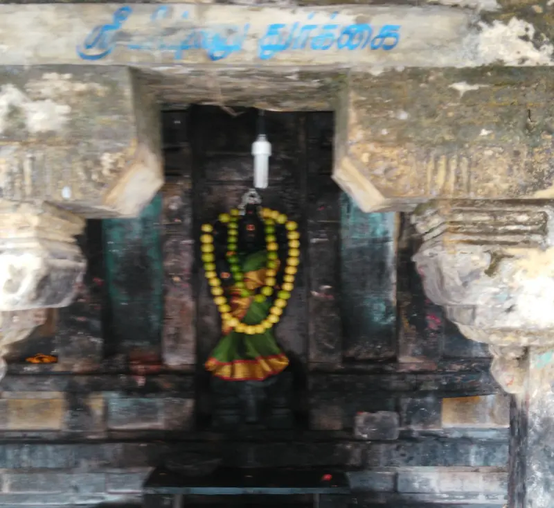 A dark idol in a green and red cloth and a yellow garland in a stone doorway; a blue Tamil name board is above.