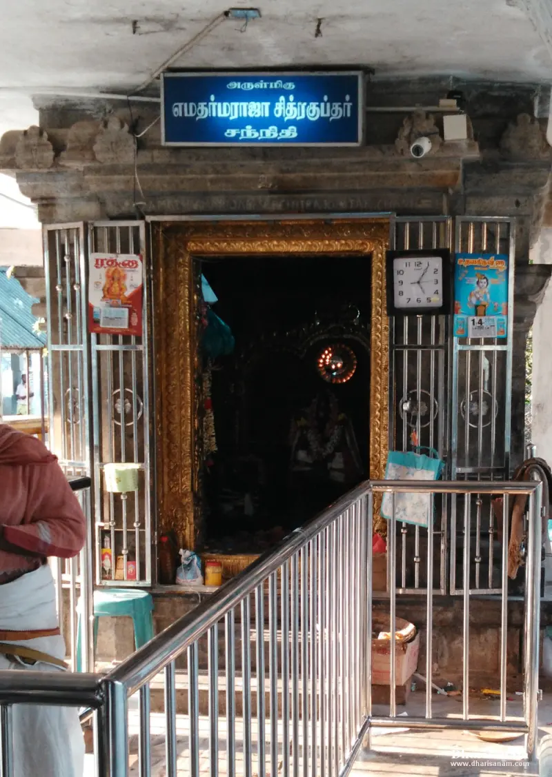 A shrine doorway with a gold frame and a blue name board, a railing in front and a devotee at left