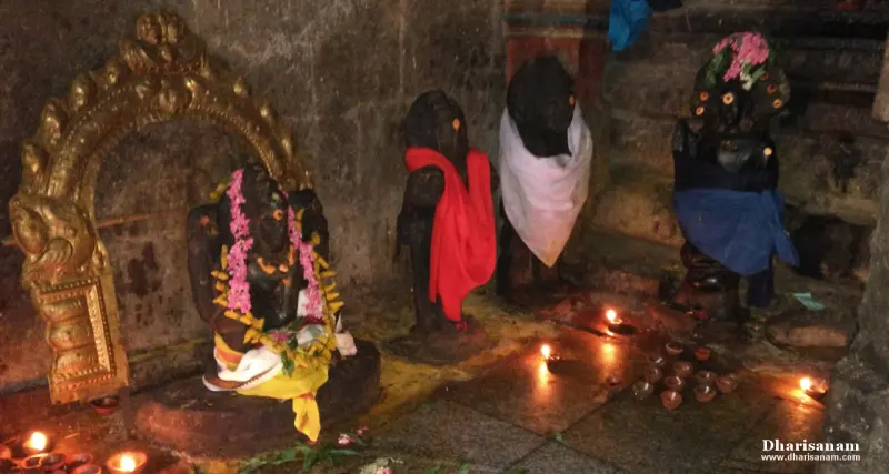 Four dark stone idols, two in red and white cloth and one garlanded, with small lamps burning at their feet