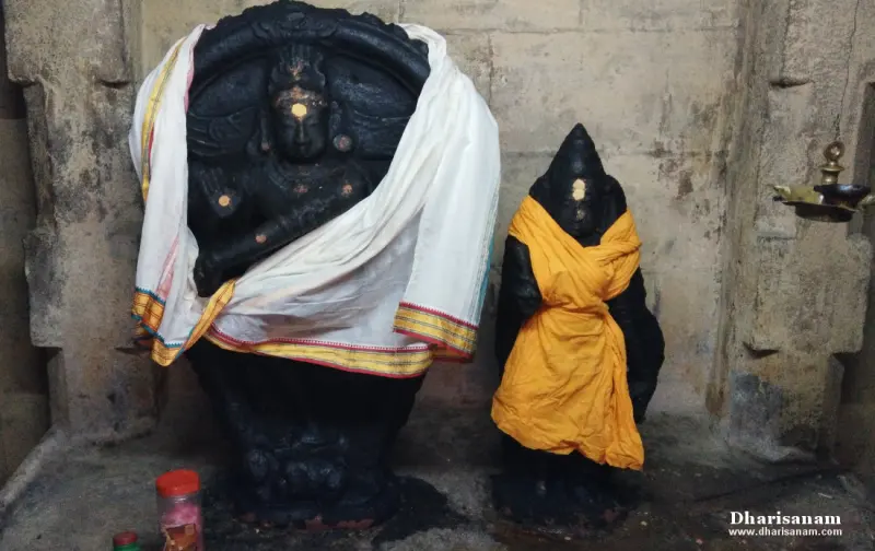 A large black idol draped in white cloth beside a smaller one in yellow cloth, with a hanging lamp at right