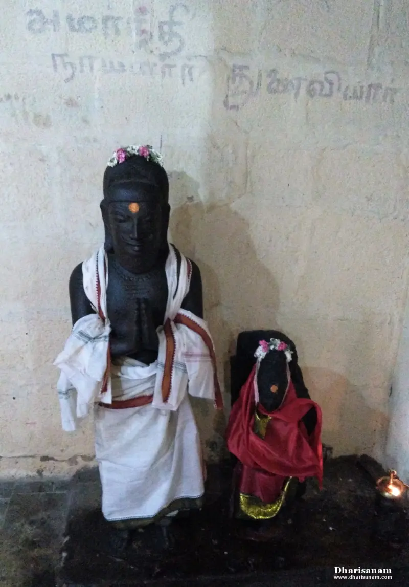 A black stone figure in a white cloth beside a smaller figure in red, in front of a wall with Tamil writing