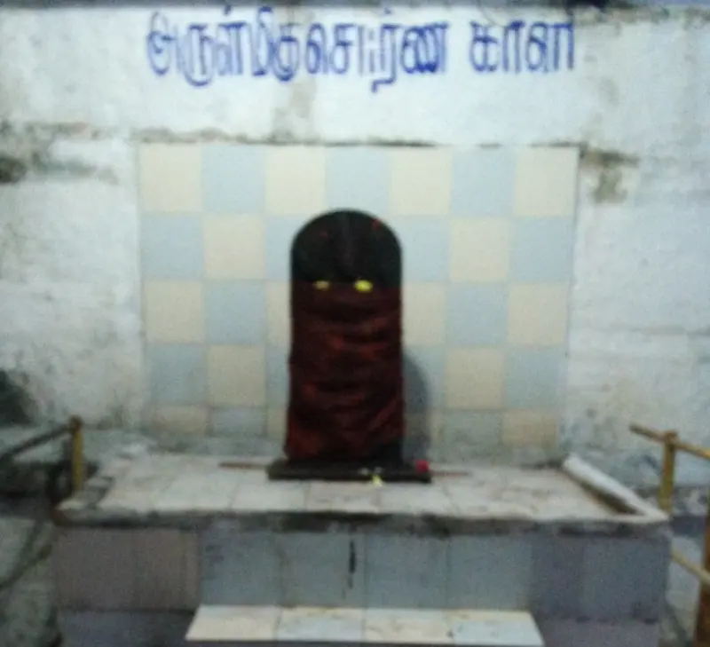 A dark figure draped in red cloth with yellow eyes on a raised platform, with a Tamil name painted on the wall above.