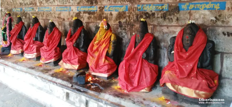 Black stone idols in a row along a wall, draped in red and gold cloth, with Tamil name boards above