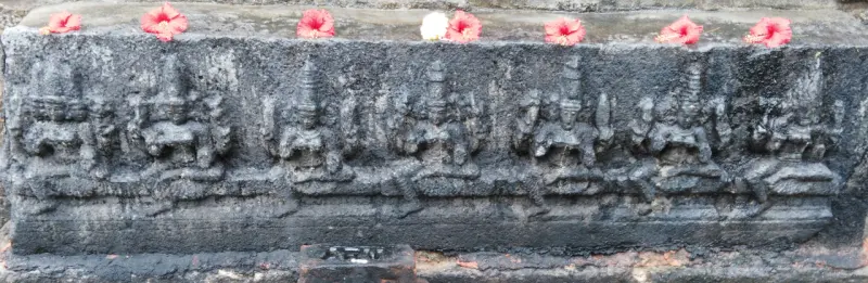 A long dark stone panel carved with a row of seated figures, with red and white flowers laid along the top.