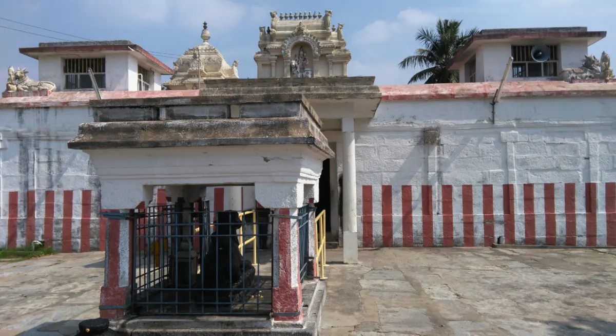A small shrine behind iron railings in a courtyard, with a painted wall striped red and white and rooftop towers behind