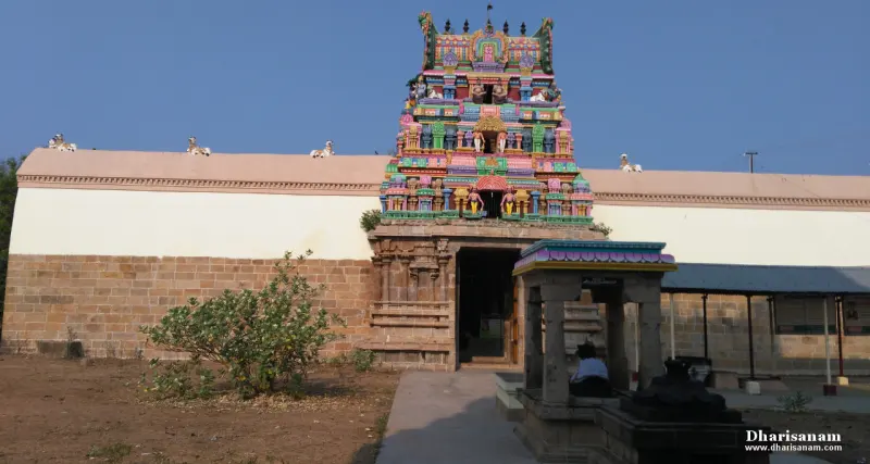 Painted gateway tower above a white wall, with a small mandapam and a seated person in front