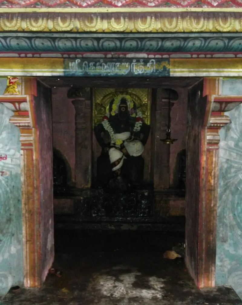 A small shrine with a seated dark idol garlanded with flowers, under a painted lintel with a Tamil name board.
