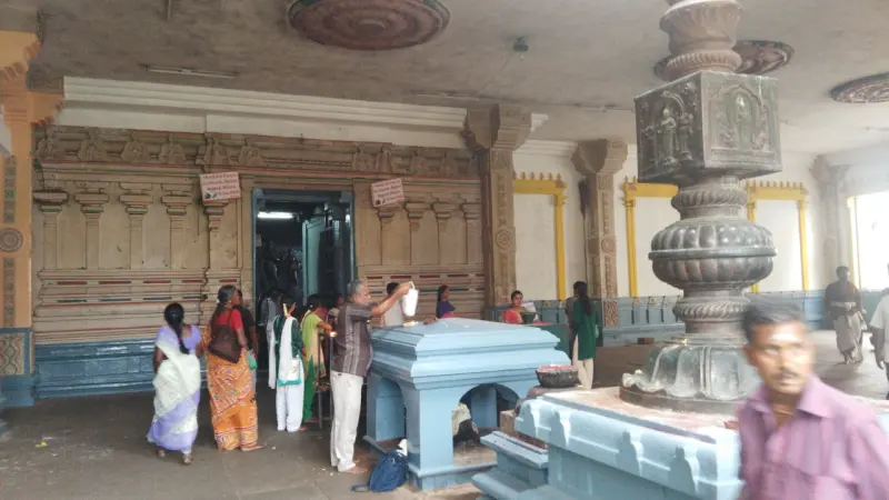 Devotees stand before a shrine doorway; a carved pillar and a small blue platform are in front.