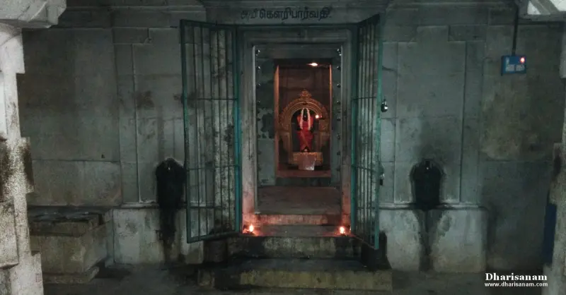 A barred doorway with a Tamil name board, and a lit idol in red cloth within.