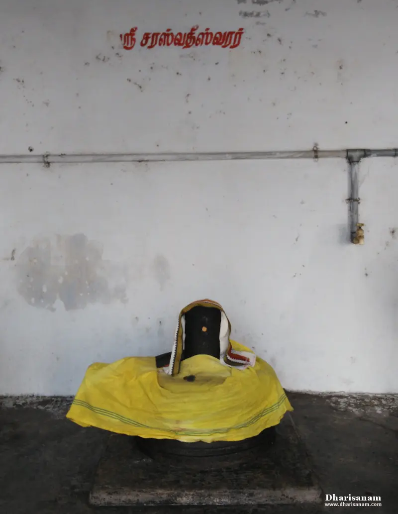 A lingam covered with yellow and white cloth on the floor, with a Tamil name painted on the wall behind it