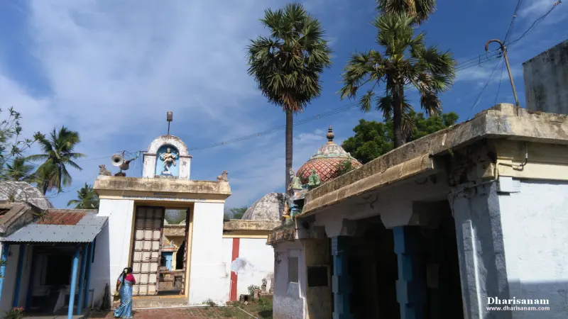 Small white shrine and low buildings among palm trees, with a woman in a blue sari standing at the left