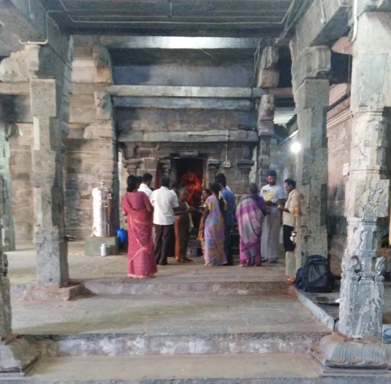 Devotees stand on stone steps in a pillared hall facing a lit doorway at the far end.