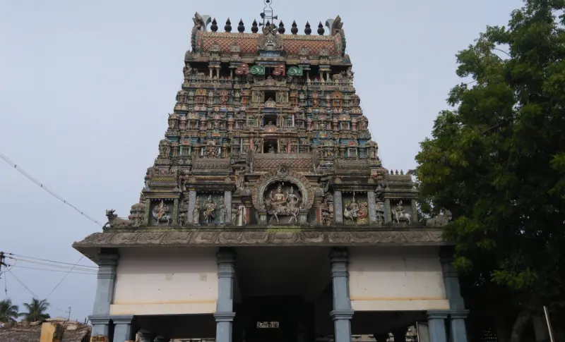 A tiered gopuram covered in colourful sculptures, with a large tree at its right.