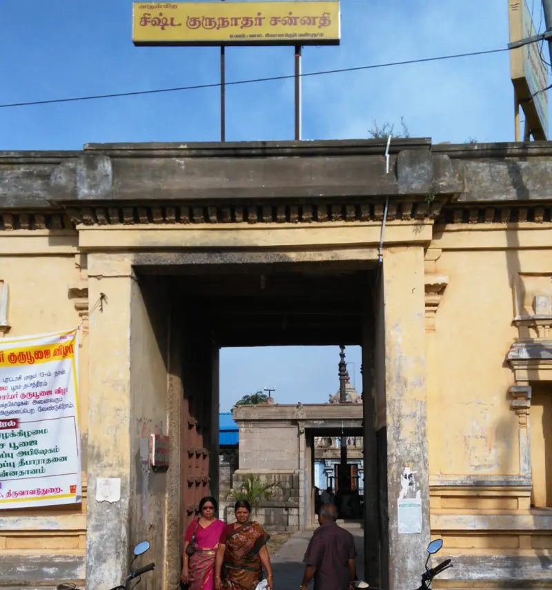 A tall stone gateway with a yellow Tamil signboard on top and three people walking in front of it