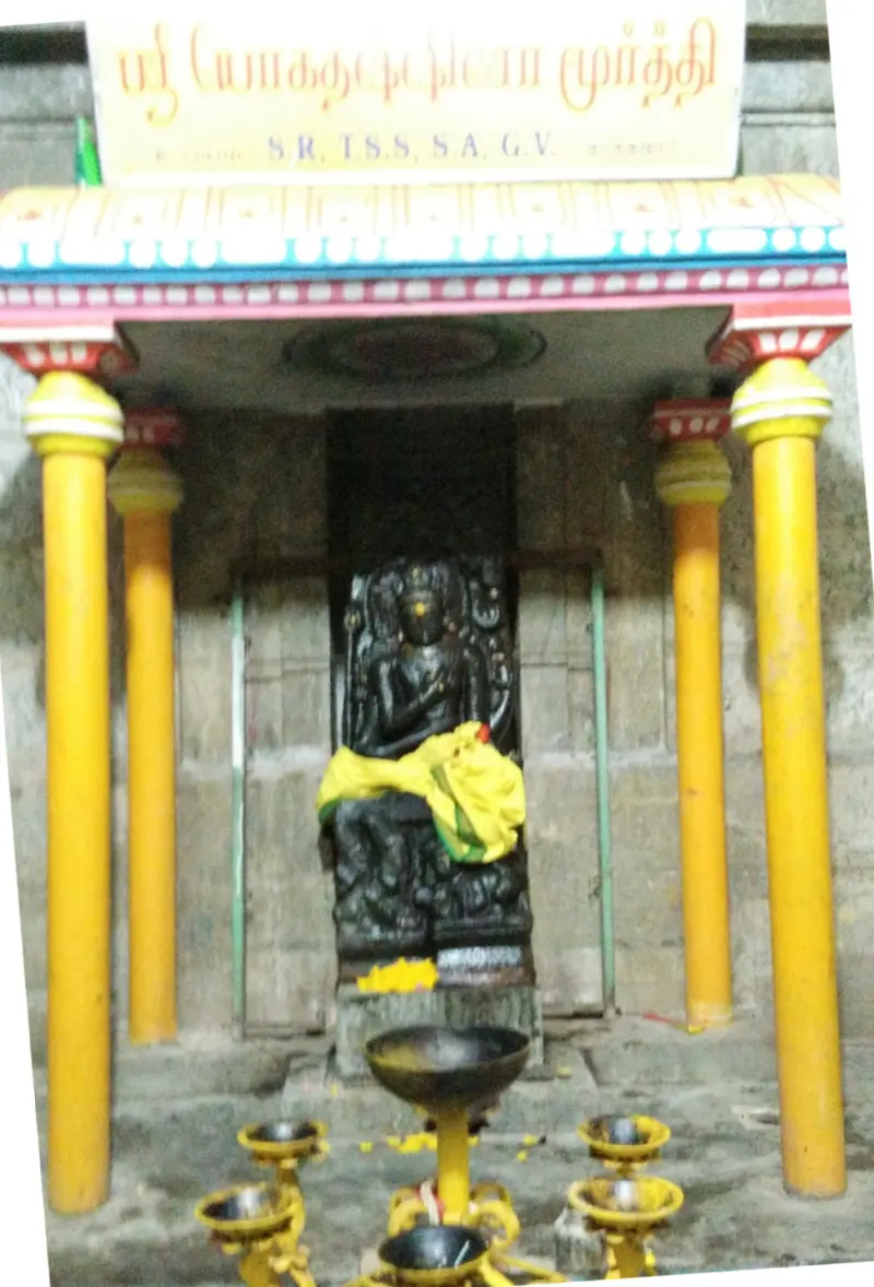 A dark stone figure seated in a niche under a canopy with yellow pillars, with lamps in front.