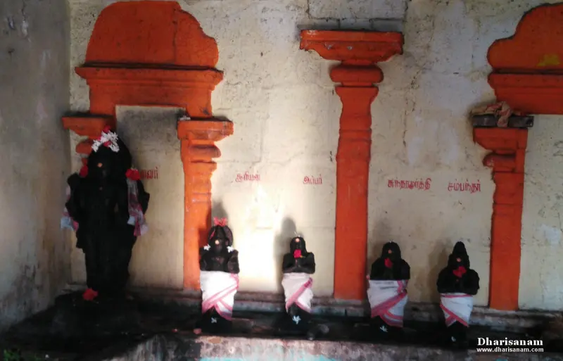 Five dark stone idols with cloth wraps in front of a wall with orange arches and red Tamil names