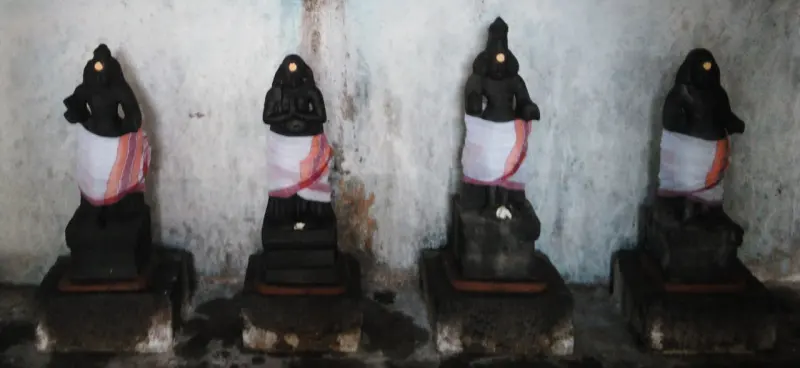 Four small black stone figures in white cloths stand in a row against a plain wall.