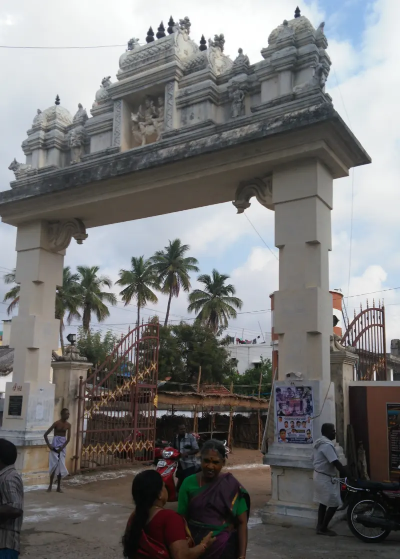 A broad stone entrance arch with carved figures on top, an open iron gate and coconut palms beyond