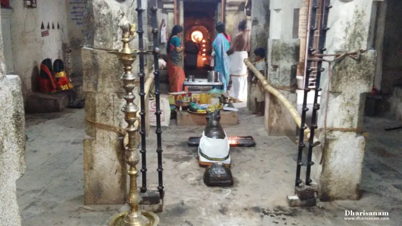 Devotees stand in a stone hall with a brass lamp stand in the front and a lit sanctum doorway behind