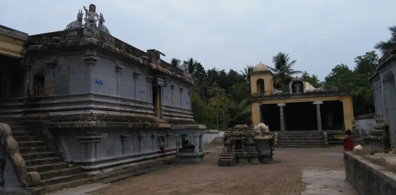 A grey stone wall with steps at the left, small shrines in the paved courtyard and a yellow porch beyond.
