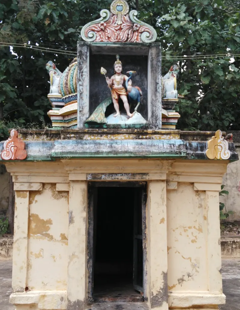 A small yellow shrine with a painted Murugan figure holding a spear, with a peacock beside him, above a dark doorway.