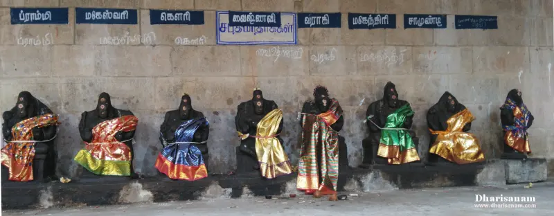 A row of dark stone idols in bright silk cloths below a wall with name boards for each in Tamil