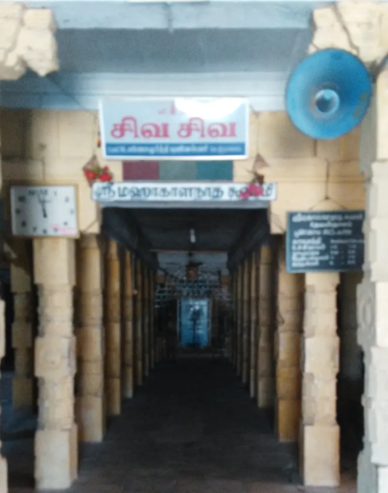 A long pillared corridor with name boards and a wall clock, leading to a lit doorway in the distance.
