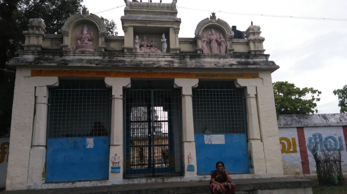 A low entrance building with a gated doorway and pink figures of deities on its upper parapet; a woman sits in front.