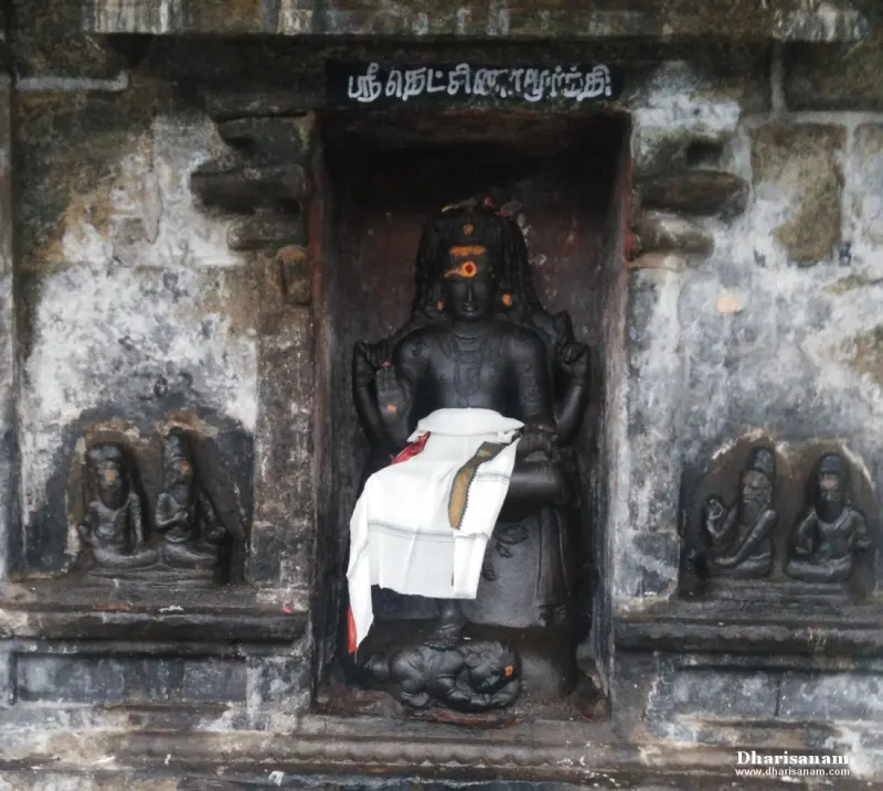 Black stone Dakshinamurthy standing in a niche under a name board, with small carved sages on both sides