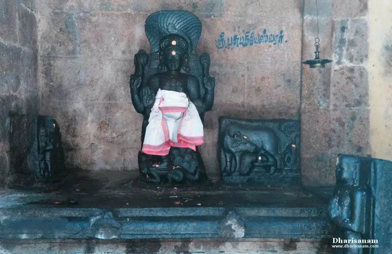 A dark stone seated idol in a white cloth, with a carved cow relief on the right and a Tamil label above