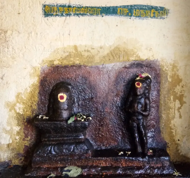 A lingam on a stone base beside a dark standing figure, under a green Tamil name board