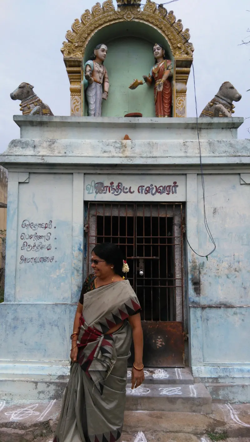 A pale blue shrine with a barred door, two Nandi figures and two standing figures above; a woman stands before it.