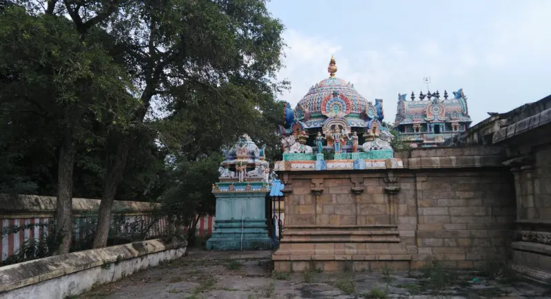 A small shrine, a domed shrine roof and a tower beyond, beside trees and a striped wall.