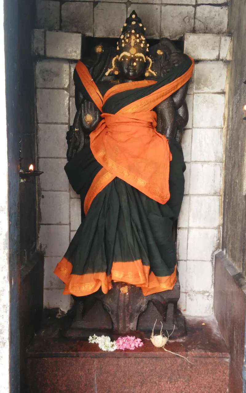 A dark stone standing figure in a wall niche, draped in orange and black cloth, flowers at its feet