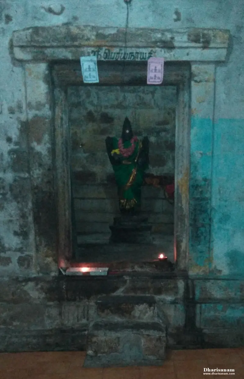 A small niche with a dark stone image in a green sari and flower garland, with a lamp before it.