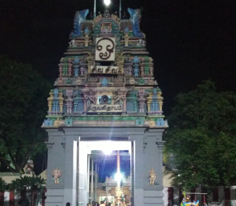 A tiered painted tower over a grey gateway, with a lit passage and crowd inside leading to the temple