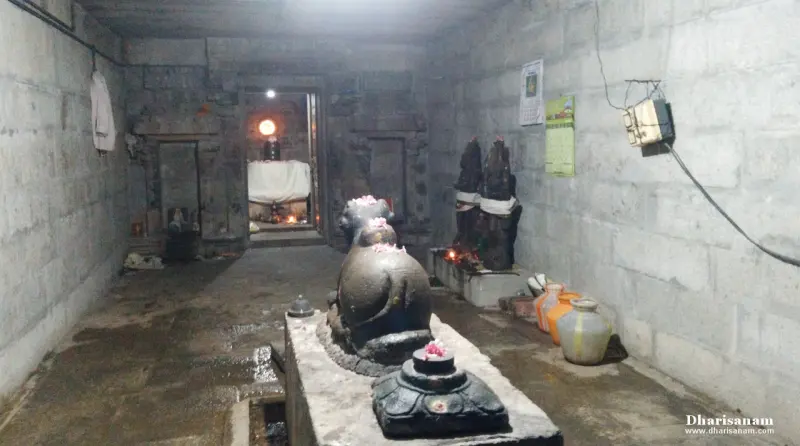 A stone Nandhi and a Balipeetam on a platform, facing the lit sanctum at the end of a dim corridor.