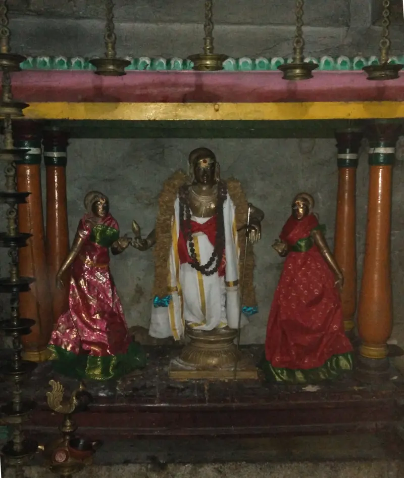 Three bronze figures in a pillared frame: a man in white with a rosary and a staff, flanked by two women in red saris.