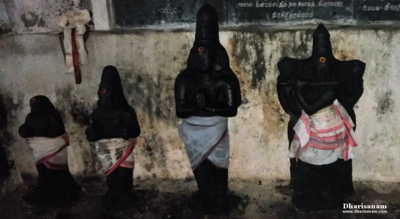 Four dark stone idols stand against a wall under a Tamil name board, two with folded hands