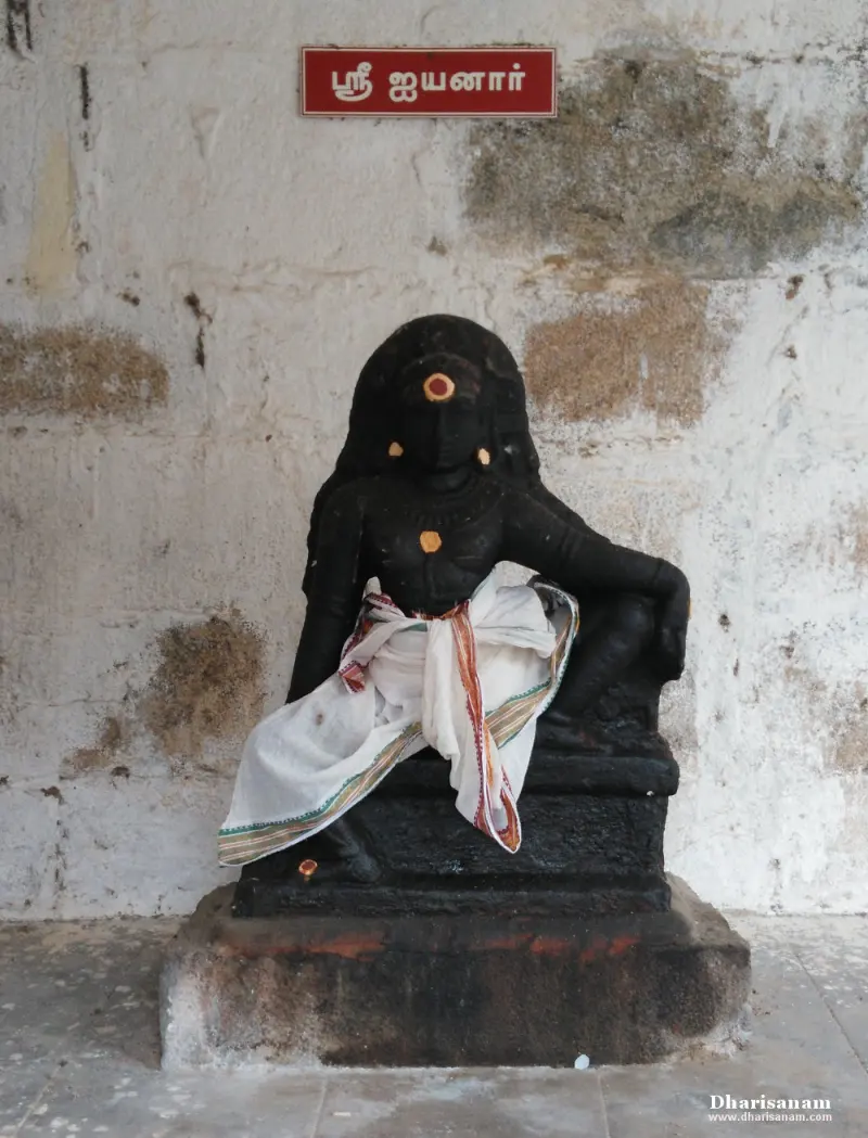A seated dark stone idol in a white cloth, with a red board above reading Sri Iyyanar in Tamil