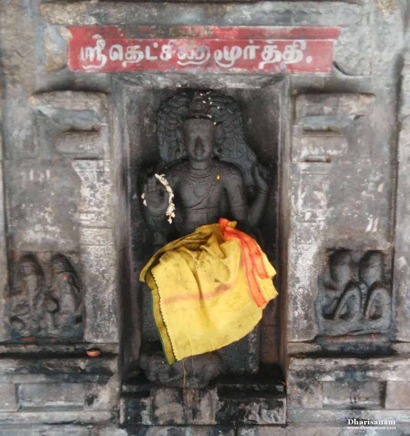 A black stone figure in a wall niche, a yellow cloth over its lap, with a red Tamil name board above