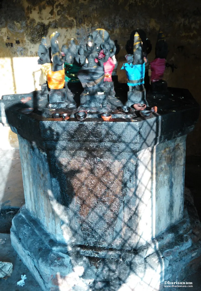 Several small dark idols in coloured cloth on a tall octagonal stone platform with clay lamps at the edge