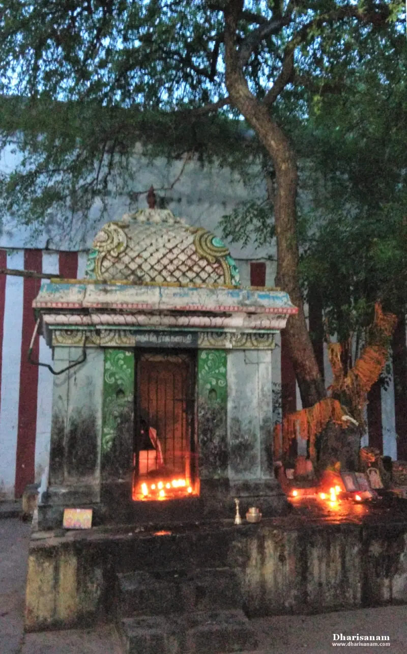 A small stone shrine with a domed roof beneath a tree, with oil lamps burning at its barred doorway.