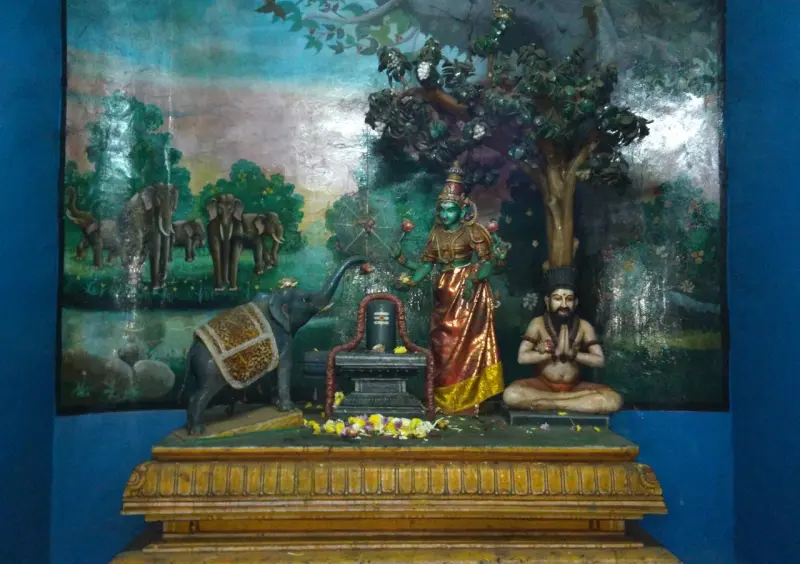 A lingam on a stand with a stone elephant, a standing goddess and a seated sage in front of a forest painting.