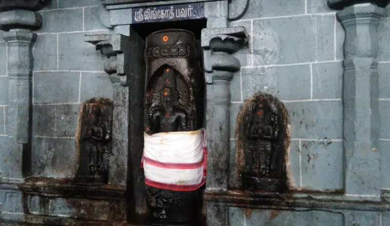 A dark stone Lingothbhavar in a niche with a Tamil name board, flanked by two carved figures in the grey stone wall.