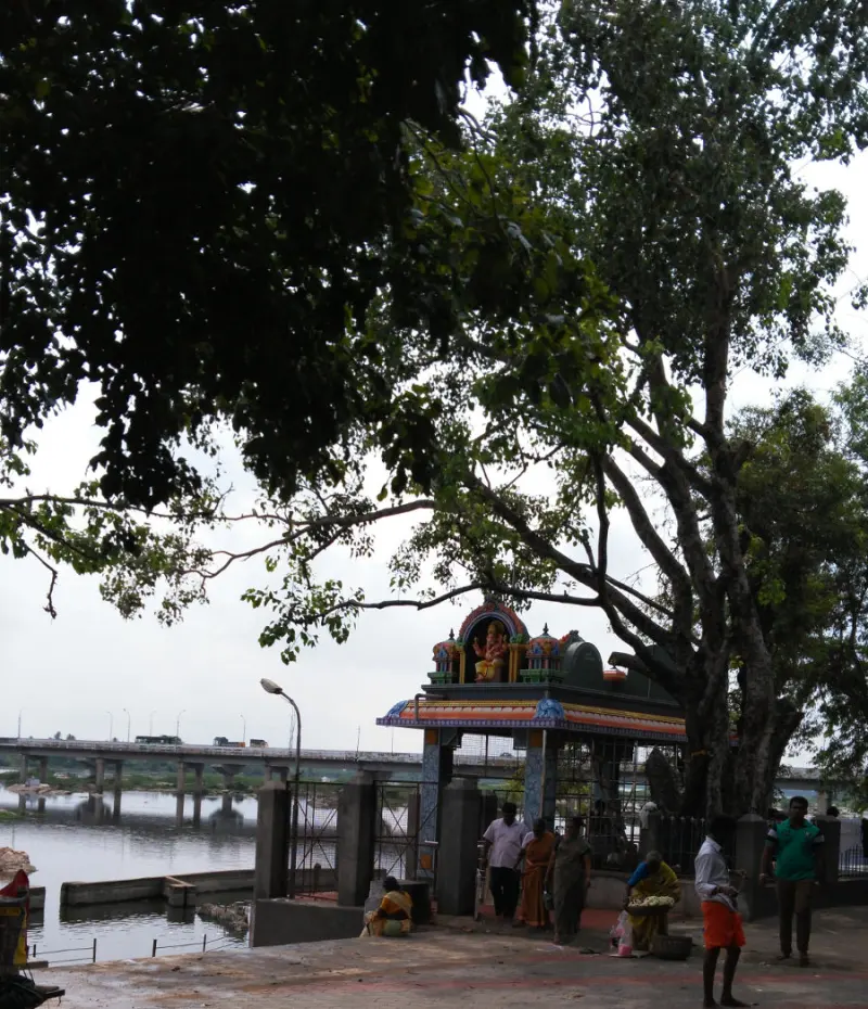 A small painted shrine under a large tree on the riverbank, with devotees standing near it and a bridge behind.