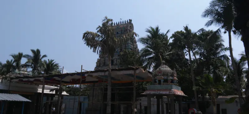 A tall gopuram rising behind palm trees and a canopy, with a small domed shrine at right.