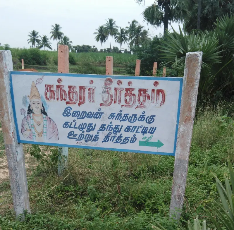 Tamil sign board for Sundarar Theertham with an arrow, set beside green fields
